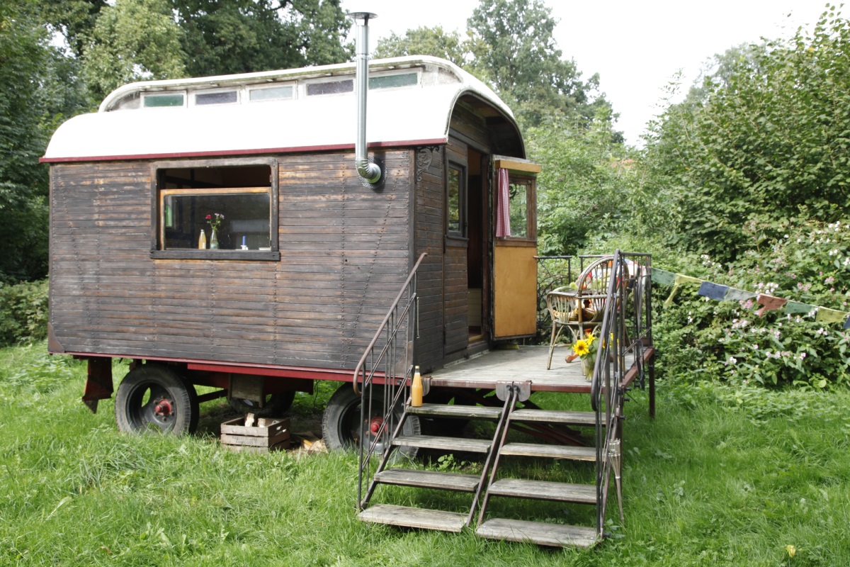Dunkelbrauner Zirkuswagen mit weißem Dach, Schornstein und Holzveranda mit Treppe