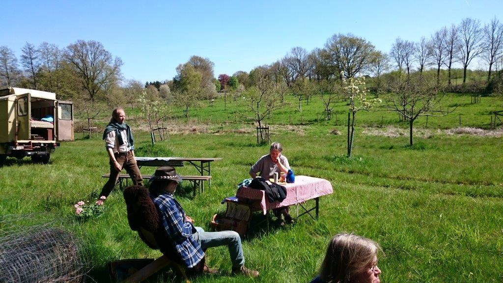Gäste sitzen an einem gedeckten Tisch auf der Streuobstwiese, Wagen im Hintergrund